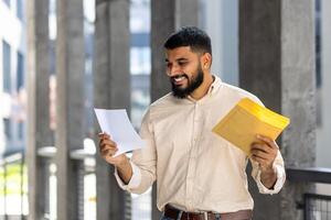 gelukkig jong Indisch Mens staand buiten gebouw, Holding envelop en glimlachend lezing ontvangen brief en resultaten foto