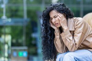 volwassen volwassen vrouw depressief en met hoofdpijn zittend Aan bank buiten kantoor gebouw, overwerkt en overwerkt bedrijf vrouw verdrietig, na werk. foto