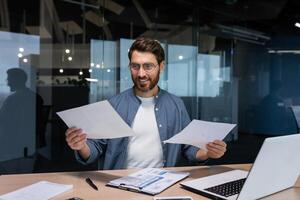 tevreden met de resultaat financier zakenman houdt in zijn handen en onderzoekt contracten en rapporten glimlachen en Vrolijk, Mens in een gewoontjes overhemd werken binnen de kantoor gebruik makend van een laptop in papierwerk. foto