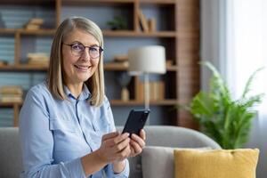 een vrolijk volwassen vrouw zittend Aan de bank, innemend met haar telefoon, en genieten van technologie in een knus huis instelling. foto