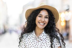 portret van een blij latina zakenvrouw met gekruld haar- en een hoed, Aan een stad straat met een vervagen achtergrond van stad lichten. foto