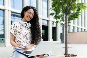 een jong spaans vrouw leerling glimlacht terwijl aan het studeren buitenshuis. ze is gezeten met een laptop en notitieboekje, uitstralend geluk en een leergierig houding in een stedelijk omgeving. foto