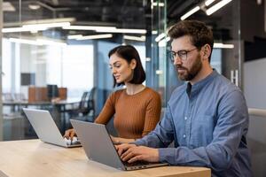 gefocust Mens en vrouw gebruik makend van laptops Bij een houten tafel in een helder, hedendaags werkruimte, belichamen professionaliteit en teamwerk. foto