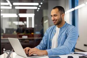 glimlachen jong moslim Mens zakenman, ontwerper, freelancer, programmeur in blauw overhemd werken in kantoor met laptop, zittend Bij kantoor tafel en typen Aan toetsenbord. foto