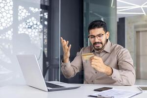 perplex volwassen mannetje in kantoor instelling onderzoeken credit kaart met een frons, wijzend op potentieel fraude of vergissing. foto