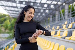 blij spaans vrouw in atletisch slijtage controle haar geschiktheid tracker na een training Bij de stadion. foto