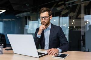 senior zakenman in bril en baard is denken over een beslissing, Mens is werken binnen de kantoor en gebruik makend van laptop Bij werk, volwassen investeerder in een bedrijf pak is zittend Bij de tafel foto