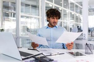 nadenkend echt zakenman lezing financieel rapport, spaans zakenman Holding document in handen op zoek teleurgesteld, werken binnen modern kantoor met laptop achter papier werk. foto