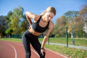 een fit vrouw in sport- kleding pauzeert met handen Aan knieën, uitgeput na een vermoeiend rennen Aan een zonnig spoor. foto