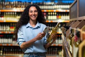 portret van gelukkig en glimlachen vrouw klant in supermarkt, Latijns Amerikaans vrouw kiezen alcohol wijn gebruik makend van app Aan telefoon naar scannen etiketten en tarief wijn, op zoek Bij camera met glimlach foto