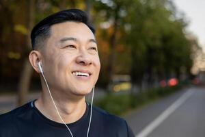 detailopname portret van een jong glimlachen Mens Aziatisch mannetje sportman staand in de midden- van de straat in koptelefoon, luisteren naar muziek, rusten, genieten van, op zoek naar de kant. foto