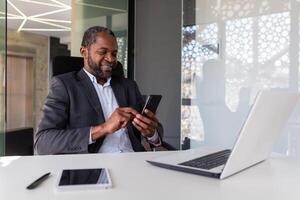 volwassen ervaren en geslaagd Afrikaanse Amerikaans baas Bij werkplaats binnen kantoor, senior tevreden met resultaten zakenman gebruik makend van telefoon, lezing nieuws van online app Aan smartphone. foto