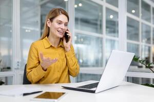 volwassen geslaagd blond bedrijf vrouw in geel overhemd gelukkig pratend Aan de telefoon met een glimlach, vrouw financier arbeider Bij de werkplaats in de midden- van de kantoor gebruik makend van een laptop Bij werk. foto