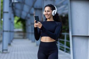 spaans sportvrouw met hoofdtelefoons luisteren naar online muziek- en audio podcasts met boeken terwijl jogging en aan het doen actief opdrachten, vrouw gebruik makend van app Aan telefoon, in trainingspak. foto