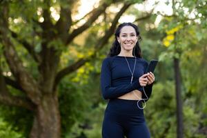 een atletisch spaans vrouw glimlachen en Holding een smartphone met oortelefoons, nemen een breken van haar rennen in een weelderig groen park. foto