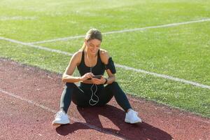 een vrouw atleet in sportkleding geniet een breken Aan de bijhouden veld, glimlachen net zo ze toepassingen haar telefoon met oortelefoons. foto