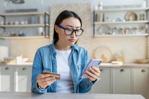 van streek Aziatisch vrouw proberen naar maken een aankoop in een online online op te slaan, vrouw Bij huis zittend Aan een sofa in de keuken gebruik makend van een toepassing Aan een smartphone en Holding een bank credit kaart foto