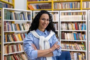 portret van een zelfverzekerd, glimlachen vrouw bibliothecaris staand met armen gekruiste in een helder gekleurde bibliotheek vol van boeken. ze draagt bril en een gewoontjes kleding. foto