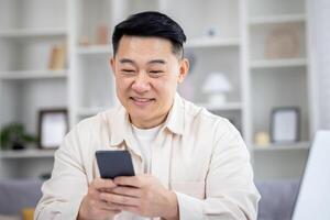 volwassen Aziatisch Mens zittend Bij tafel Bij huis, Mens gebruik makend van telefoon, typen bericht Aan smartphone, app gebruiker en browsen internet in huis bioscoop kamer, glimlachen tevreden. foto