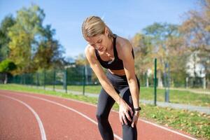 fit vrouw in atletisch slijtage rust met handen Aan knieën na rennen Aan bijhouden veld, weergeven training vermoeidheid en bepaling. foto