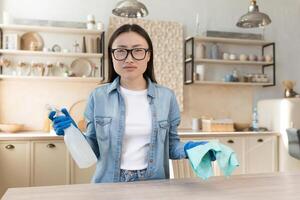 portret van ontevreden jong Aziatisch vrouw, huisvrouw in bril op zoek Bij camera Holding wasmiddel en spons foto