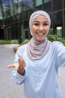 vrolijk moslim vrouw met hijab vangt een selfie met een smartphone, glimlachen blij in een modern stad omgeving. foto