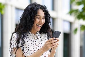 blij vrouw met gekruld haar- lachend terwijl op zoek Bij smartphone, buitenshuis met wazig achtergrond, overbrengen geluk en digitaal levensstijl. foto