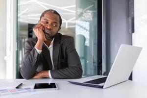 verveeld zakenman werken Aan saai werk project binnen kantoor met laptop, Afrikaanse Amerikaans volwassen volwassen Mens zittend Bij bureau. foto