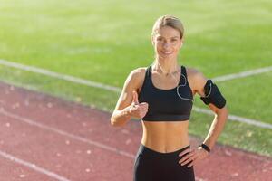 fit vrouw in sportkleding met armband en oortelefoons Aan rennen bijhouden geven duimen omhoog, uitstralend positiviteit. foto