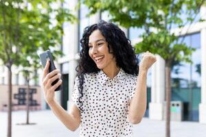 jong blij vrouw winnaar ontvangen online kennisgeving Aan telefoon, spaans vrouw met gekruld haar- vieren succes en triomf wandelen in stad in de buurt kantoor gebouw buiten. foto