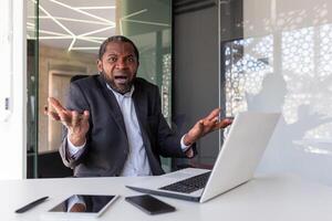 gefrustreerd boos baas op zoek Bij camera, senior grijs haren volwassen Mens geschreeuw van streek, zakenman binnen kantoor ongelukkig met prestatie resultaten, werken met laptop. foto