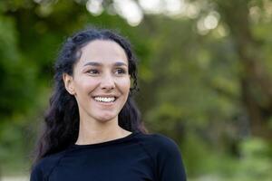 portret van een inhoud jong vrouw glimlachen voorzichtig, met groen bomen in de achtergrond, beeltenis geluk en kalmte. foto