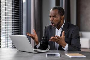 agressief zwart Mens schreeuwen in wanhoop terwijl werken door laptop in elegant appartement interieur. Afrikaanse Amerikaans mannetje arbeider geschokt door computer operationeel afbreken en systeem fout Aan apparaat. foto