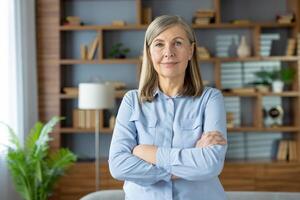 portret van een zelfverzekerd senior vrouw met armen gekruiste staand in een elegant modern leven kamer. haar sereen uitdrukking straalt uit professionaliteit en beleven. foto