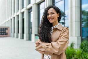 jong mooi spaans vrouw met gekruld haar- buiten kantoor gebouw, bedrijf arbeider glimlachen en op zoek Bij camera met gekruiste armen, tevreden zakenvrouw. foto