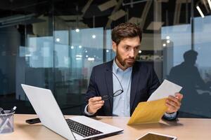 een geschokt jong Mens is zittend in de kantoor Bij de bureau, Holding een envelop met een brief en bril. kreeg slecht nieuws, rekening, credit overeenkomst. foto