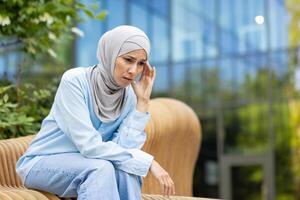een moslim vrouw in een hijab verschijnt verontrust of contemplatief terwijl zittend alleen Aan een park bank buiten. foto