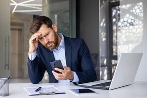 geschokt jong Mens zakenman, advocaat, bankier zittend in kantoor Bij bureau, Holding hand- naar hoofd en op zoek gefrustreerd Bij telefoon scherm. foto