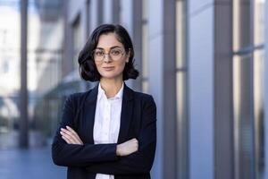 portret van een glimlachen jong vrouw advocaat staand buiten de gerechtsgebouw in een bedrijf pak, armen gevouwen aan de overkant haar borst, glimlachen en vol vertrouwen op zoek Bij de camera. foto