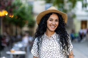 mooi jong Latijns Amerikaans vrouw portret, vrouw wandelen in avond stad in hoed met gekruld haar- in warm het weer, glimlachen en op zoek Bij camera dichtbij omhoog. foto