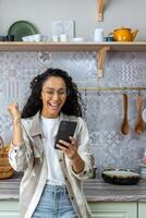 verticaal schot, Latijns Amerikaans vrouw Bij huis in keuken kreeg mooi zo nieuws online, vrouw met gekruld haar- en bril vieren zege succes Holding slim telefoon. foto