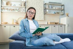 portret van een jong mooi Aziatisch vrouw in denim kleren en bril zittend Bij huis Aan de sofa Aan de achtergrond van de keuken. hij houdt een groen boek in zijn handen, leest, looks Bij de camera. foto