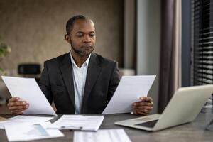 een professioneel Afrikaanse Amerikaans zakenman aandachtig beoordelingen papierwerk terwijl zittend Bij zijn bureau in een modern kantoor opstelling, tonen toewijding en concentratie. foto