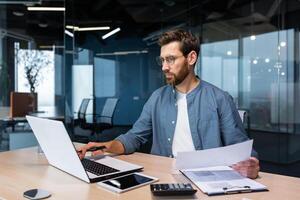 echt en gefocust financier accountant Aan papier werk binnen kantoor, volwassen Mens gebruik makend van rekenmachine en laptop voor berekenen rapporten en samenvatten rekeningen, zakenman Bij werk in gewoontjes kleren. foto