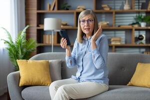 een volwassen volwassen vrouw zittend Aan een bankstel verschijnt geschokt en in ongeloof terwijl Aan een telefoon telefoontje in een knus huis instelling. foto