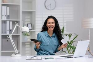 productief spaans vrouw met een blij uitdrukking beheren bedrijf taken van haar huis kantoor, Holding papieren en een rekenmachine. foto