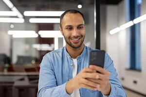 detailopname foto van een jong spaans Mens werken in een kantoor, zittend Bij een bureau, Holding een mobiel telefoon en pratend Aan een telefoongesprek.