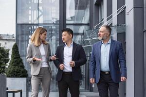 drie verschillend bedrijf mensen wandelen en pratend gefocust en attent ernstig buiten kantoor gebouw, mannetje en vrouw, bespreken plannen en werk project foto