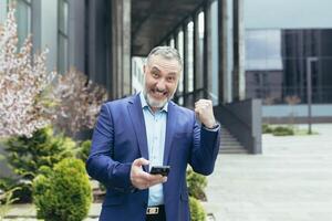 een senior grijs haar Mens in een bedrijf pak is staand in de buurt een kantoor centrum met een telefoon in zijn handen. verheugt zich, viert winnend de loterij, Toneelstukken Aan de telefoon. shows met zijn hand- ja. foto