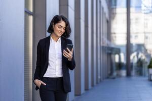 een jong mooi bedrijf vrouw is wandelen buiten de kantoor gebouw, een vrouw in bedrijf kleren is lachend, gebruik makend van een toepassing Aan de telefoon, browsen de internetten, typen een bericht. foto